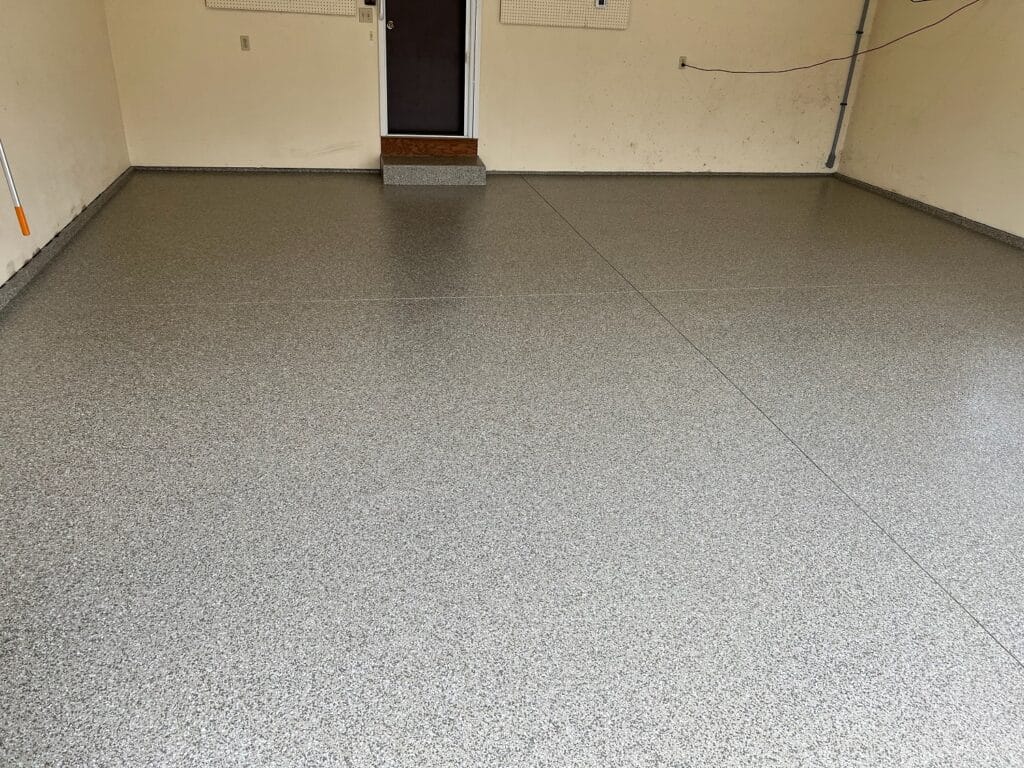 An empty room with a speckled grey epoxy floor. There's a brown door at the end, white walls with a visible electrical conduit, and no furniture.