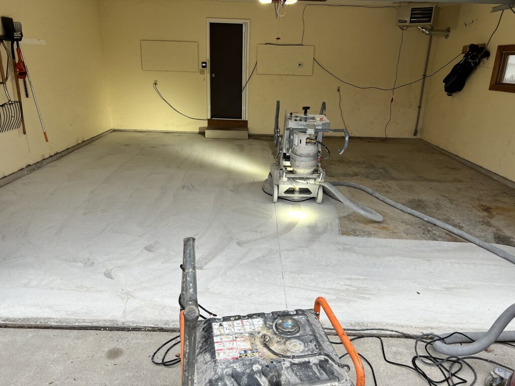 We see an empty garage with concrete flooring partially covered by a white protective sheet. Industrial floor grinding equipment is featured prominently in the foreground.