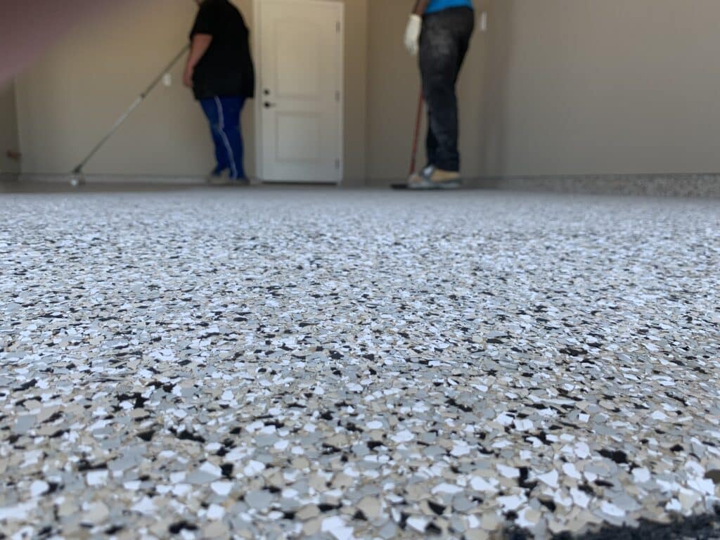 The foreground shows a speckled floor surface in focus. In the blurred background, two people are standing near a white door, one holding a broom.