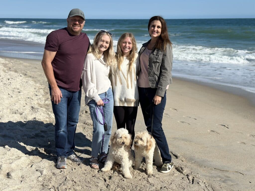 This is a photo of four people and two dogs on a sandy beach with waves in the background. They are all smiling, enjoying the sunny day.