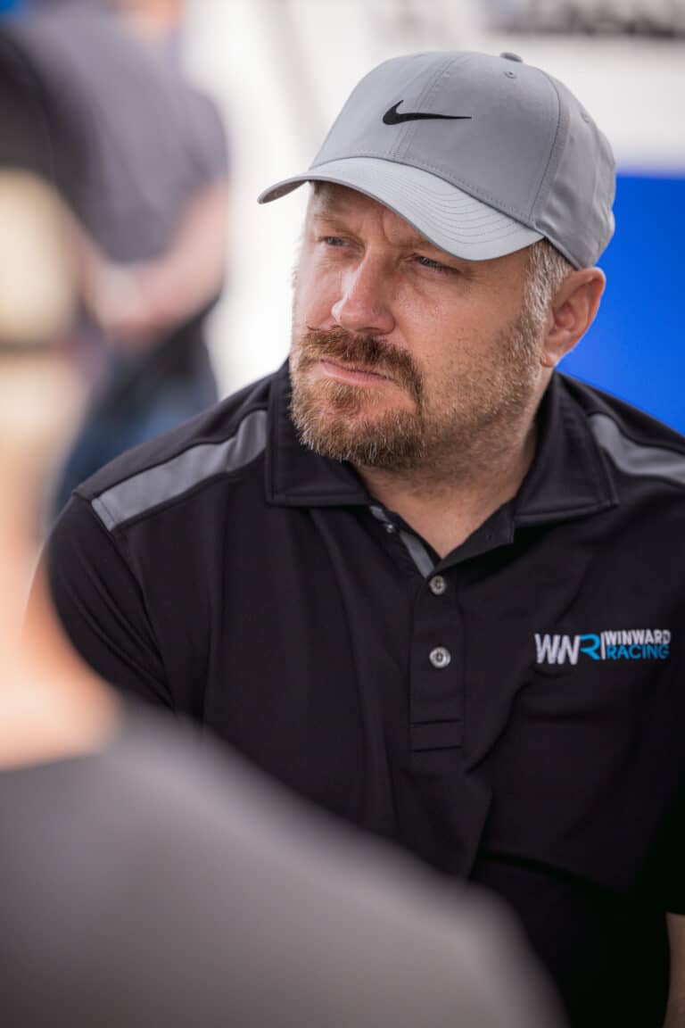 This image shows a focused person wearing a gray cap and black polo shirt with a "WINWARD RACING" logo, partially listening to someone off-camera.