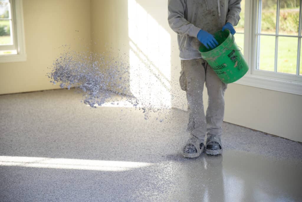 A person wearing protective gear scatters decorative chips on a freshly coated epoxy floor in a bright, sunlit room.
