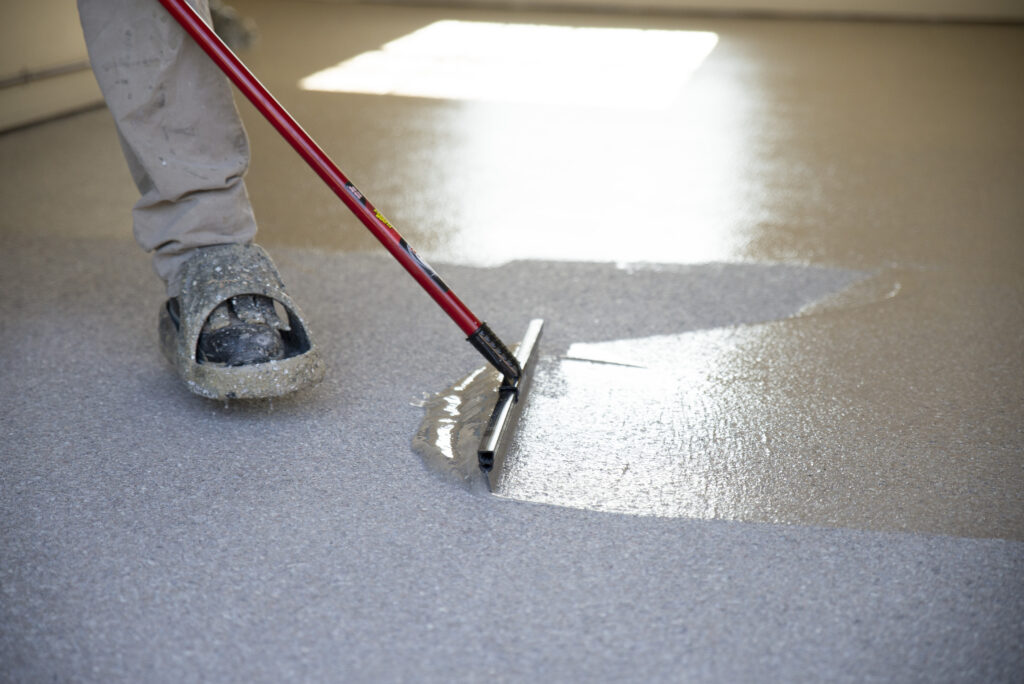 A person applies a gray coating on a floor using a squeegee. They wear protective clothing and shoes, suggesting a professional epoxy or paint application process.